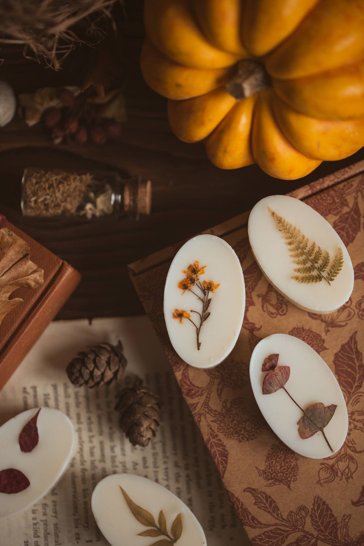 A styled flat-lay of white oval botanical wax melts decorated with pressed autumn leaves, arranged on a patterned brown paper mat alongside a yellow pumpkin, pinecones, and a small glass vial.