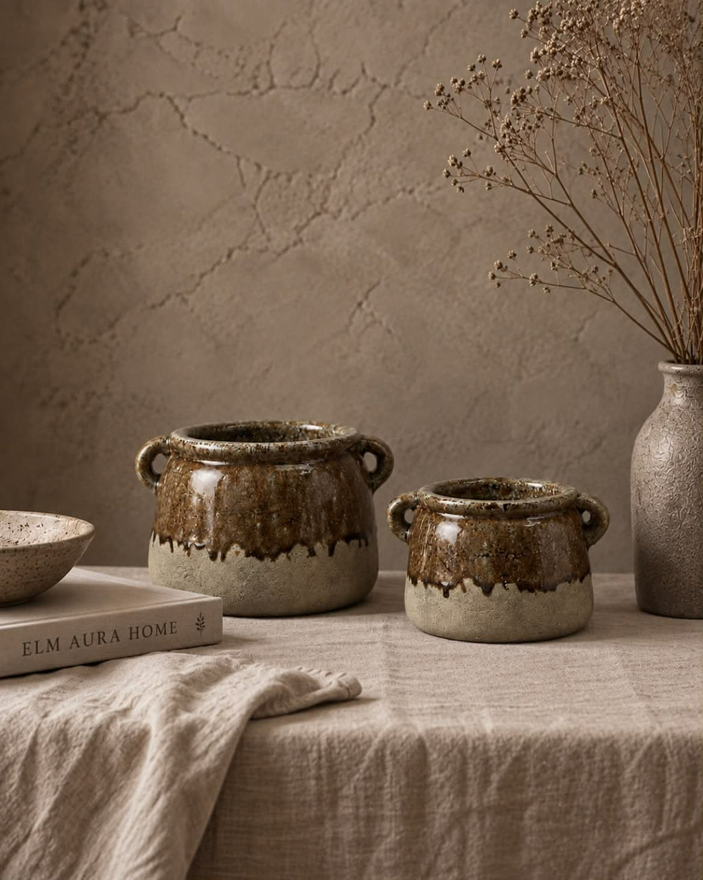 Two rustic stoneware pots with a brown and beige dripping glaze, styled on a linen-covered table with a textured stucco wall background.