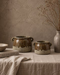 Two rustic stoneware pots with a brown and beige dripping glaze, styled on a linen-covered table with a textured stucco wall background.