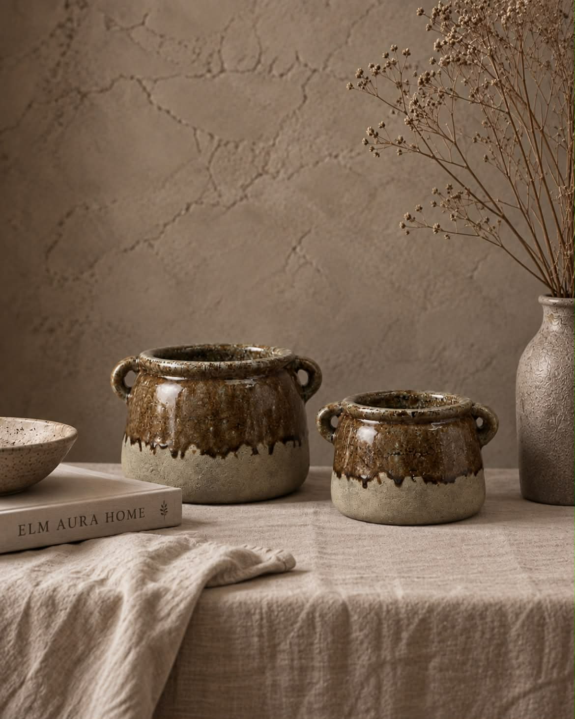 Two rustic stoneware pots with a brown and beige dripping glaze, styled on a linen-covered table with a textured stucco wall background.