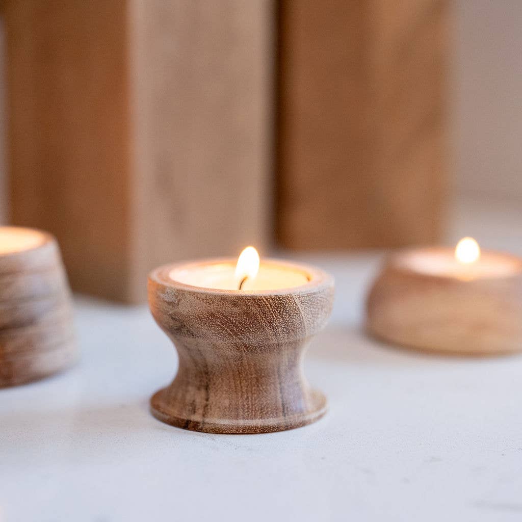 A small lit tea light in a round wooden holder sits on a light surface, with two more matching wooden candle holders blurred in the background.