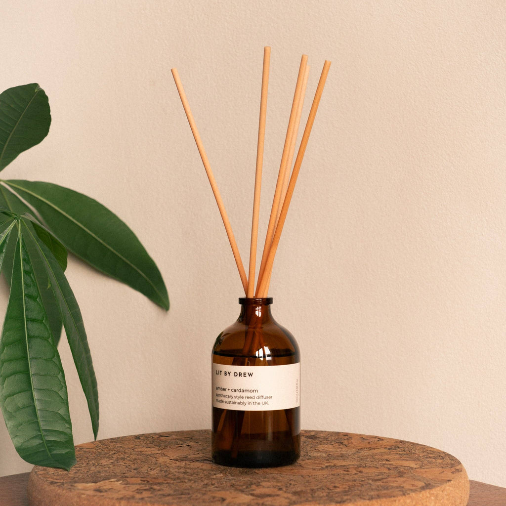 Amber glass reed diffuser with natural wood reeds on a round wooden tray next to a green leaf.