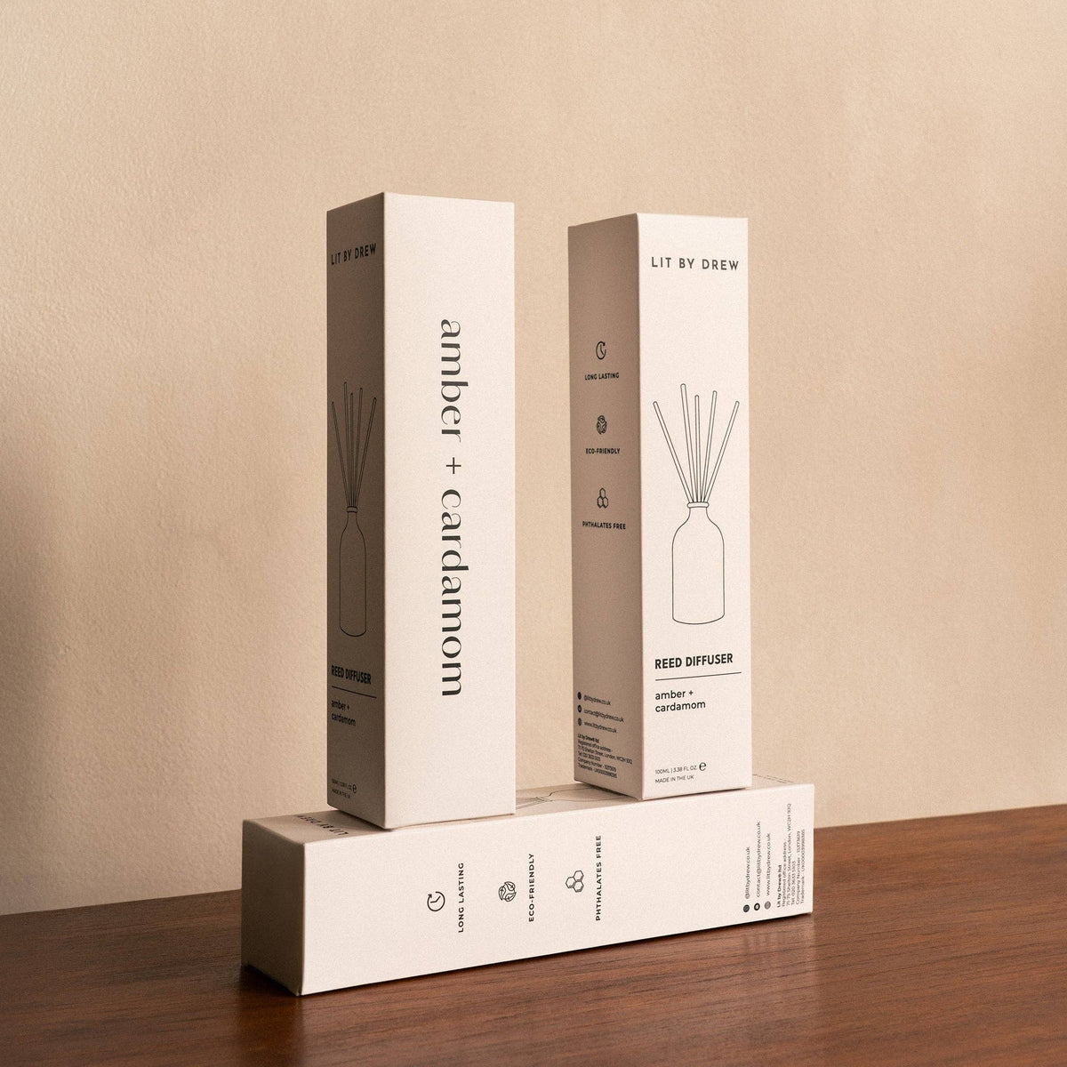 Three white Amber & Cardamom reed diffuser boxes stacked on a wooden surface against a beige wall.