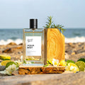 A clear rectangular perfume bottle labelled 'Aqua Mist' with a black cap, styled on a beach with ocean waves in the background, surrounded by limes, white flowers, and a wooden slice prop.
