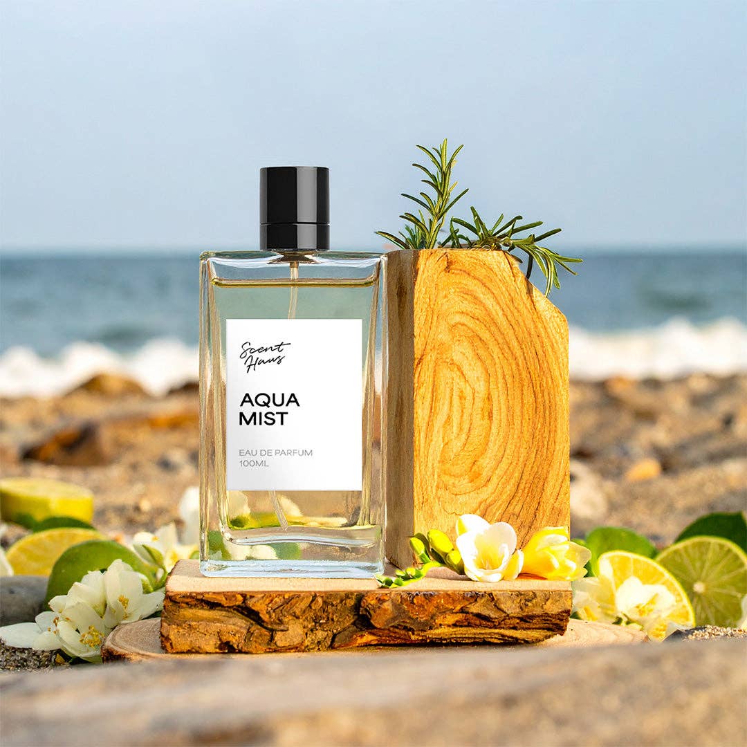 A clear rectangular perfume bottle labelled 'Aqua Mist' with a black cap, styled on a beach with ocean waves in the background, surrounded by limes, white flowers, and a wooden slice prop.