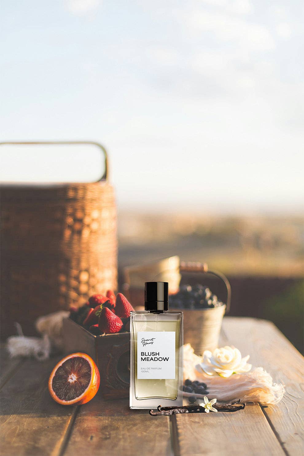 Perfume bottle labeled 'Blush Meadow' on a wooden surface with flowers and fruit.