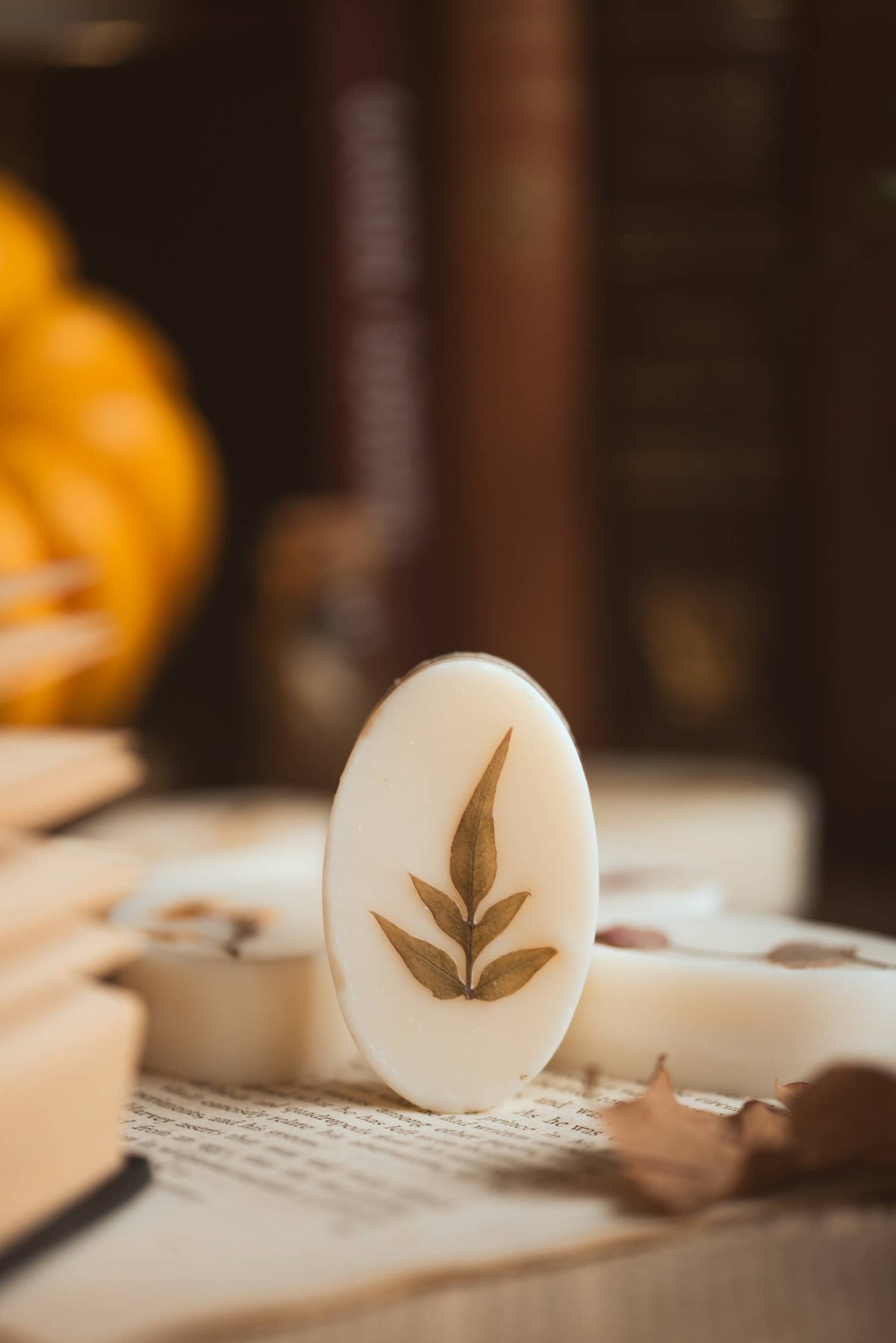An oval white botanical wax melt featuring a pressed olive leaf design standing upright on an open book page, set against a autumn background of stacked books and a pumpkin.