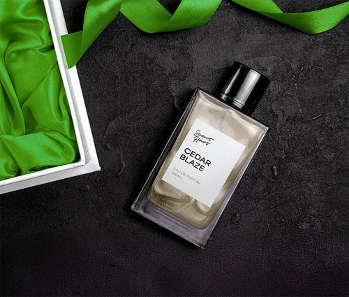 A bottle of Cedar Blaze Eau de Parfum by Scent Haus, styled on a dark textured surface with a green satin ribbon and a white gift box with green fabric lining.