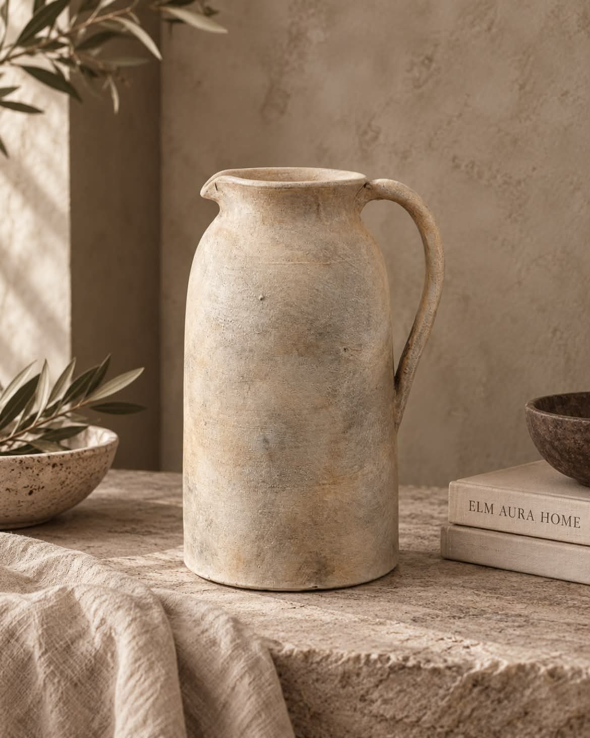 A rustic terracotta jug vase with a textured, matte finish in warm beige and cream tones, styled on a stone table with linen cloth and books.