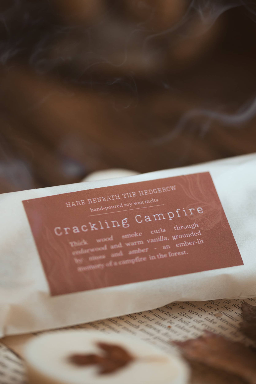 A close-up of a "Crackling Campfire" scented wax melts wrapped in off-white paper with a dusty rose-brown label, set against a rustic, smoky background.