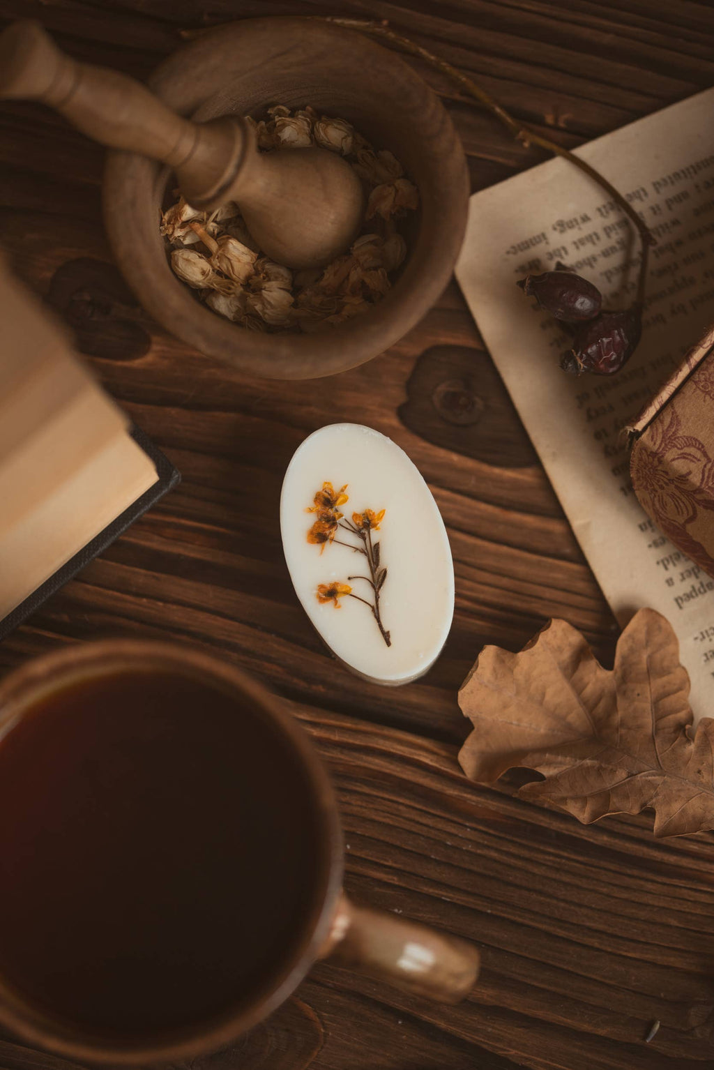 A styled flat-lay on a rustic wooden table featuring a white oval botanical wax melt topped with a dried flower, surrounded by a wooden mortar and pestle, an open book, a cup of tea, and a dried leaf.