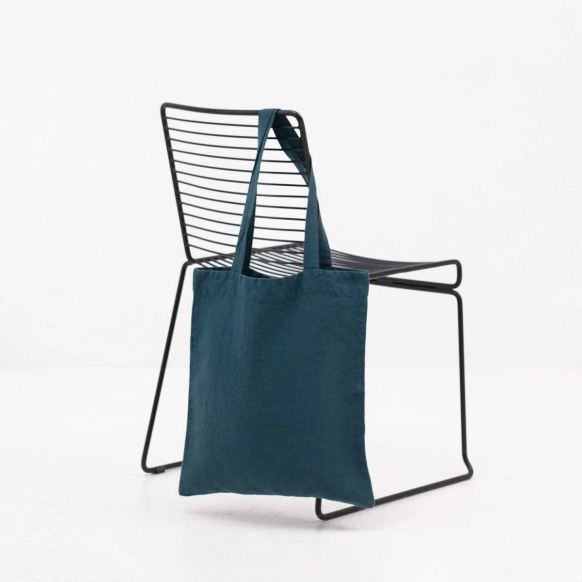 A deep water blue linen tote bag hanging over the back of a metal chair against a plain white background.