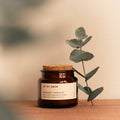 Eucalyptus and cedarwood natural scented candle in an amber glass jar with a cork lid on a wooden surface next to fresh eucalyptus.