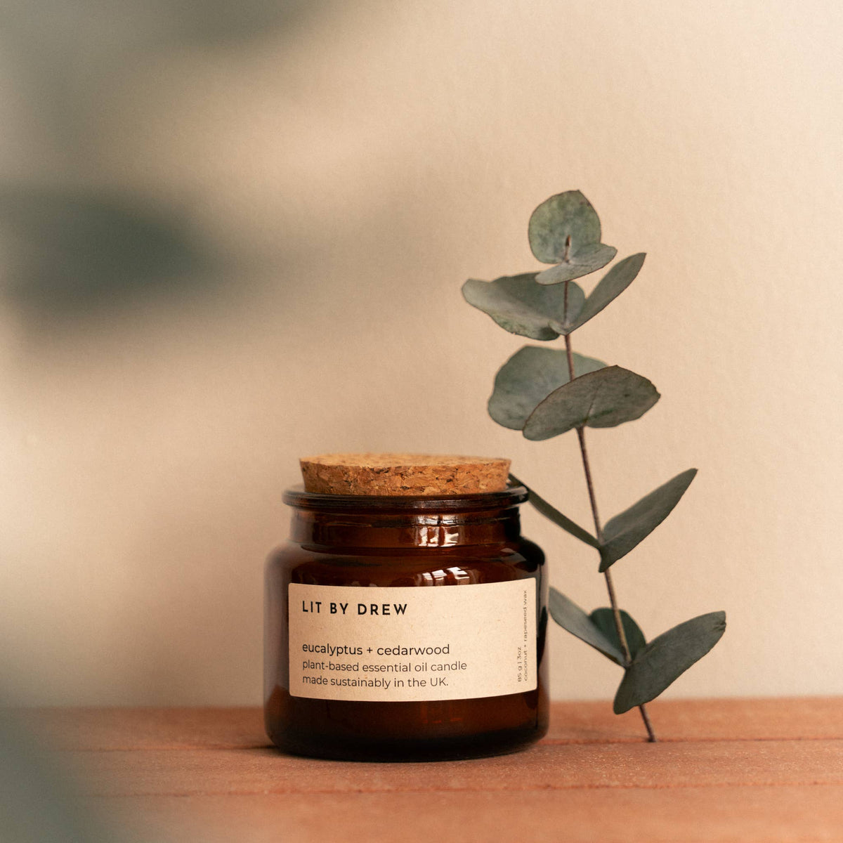 Eucalyptus and cedarwood natural scented candle in an amber glass jar with a cork lid on a wooden surface next to fresh eucalyptus.