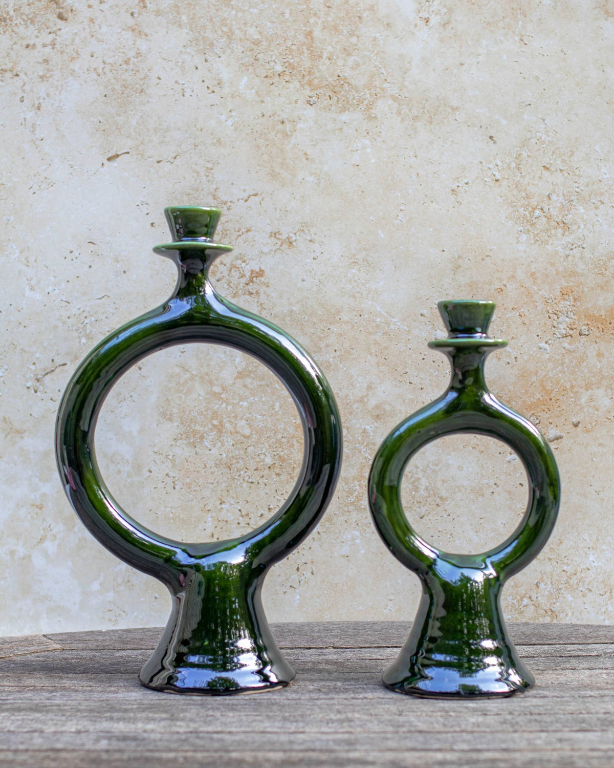 Two dark green glazed handmade Moroccan ceramic ring-shaped candle holders, displayed on a rustic wooden surface against a textured beige stone wall.