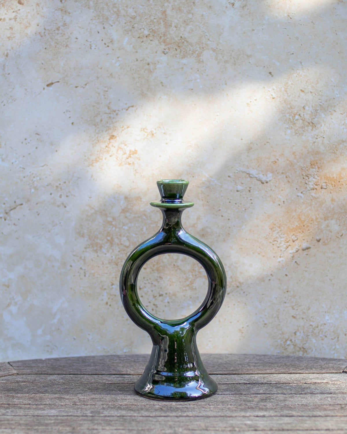 A glossy dark green handmade Moroccan ceramic candle holder with a distinctive ring-shaped body, displayed on a rustic wooden surface against a textured beige plaster wall with soft natural shadows.