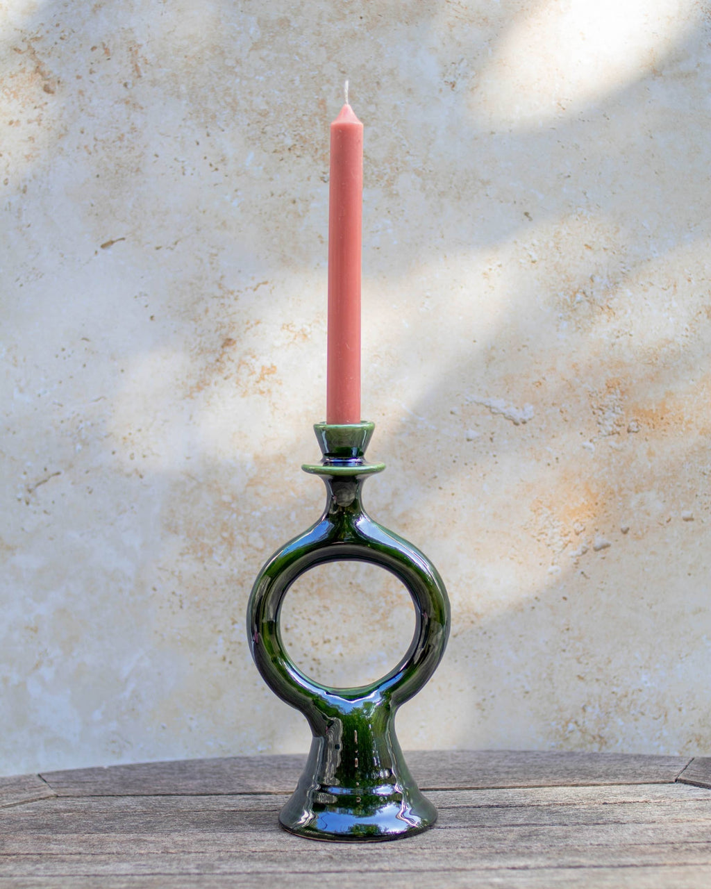 A handmade Moroccan dark green  glazed ceramic candlestick holder with a circular ring design, holding a single pink taper candle, set on a rustic wooden surface against a textured beige wall.
