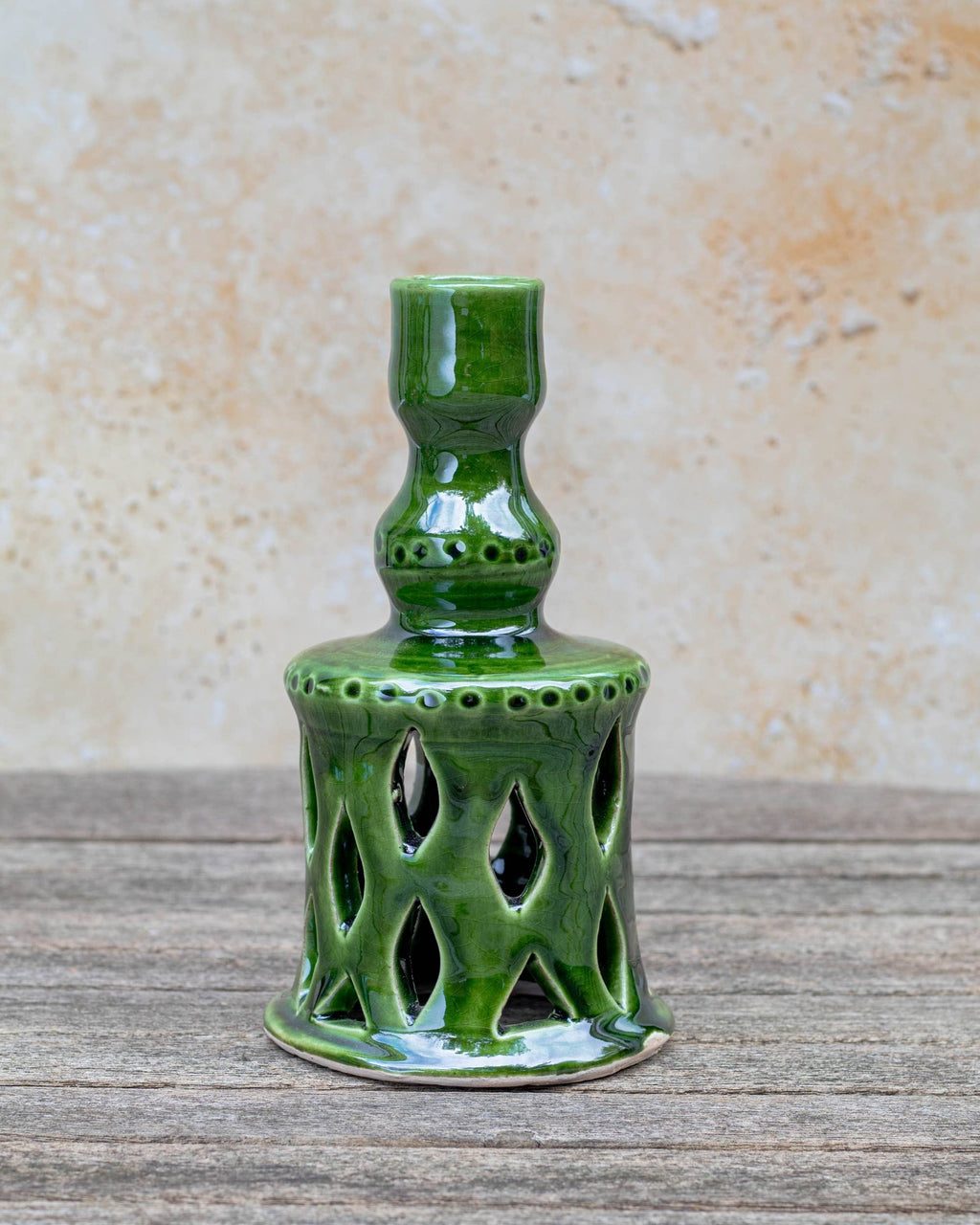 A small glossy green ceramic candlestick holder with a flared, perforated base and a narrow neck, standing on a rustic wooden surface against a textured beige wall.