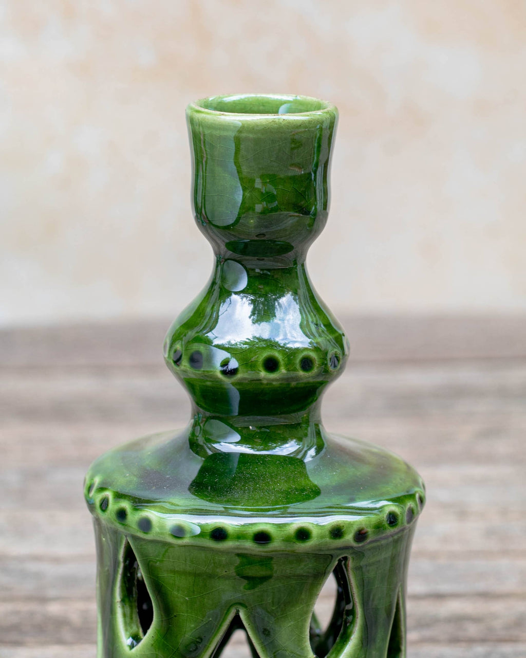 A glossy green ceramic candle holder with a flared rim and decorative cutout details on the base, photographed against a softly blurred beige background and rustic wood surface.