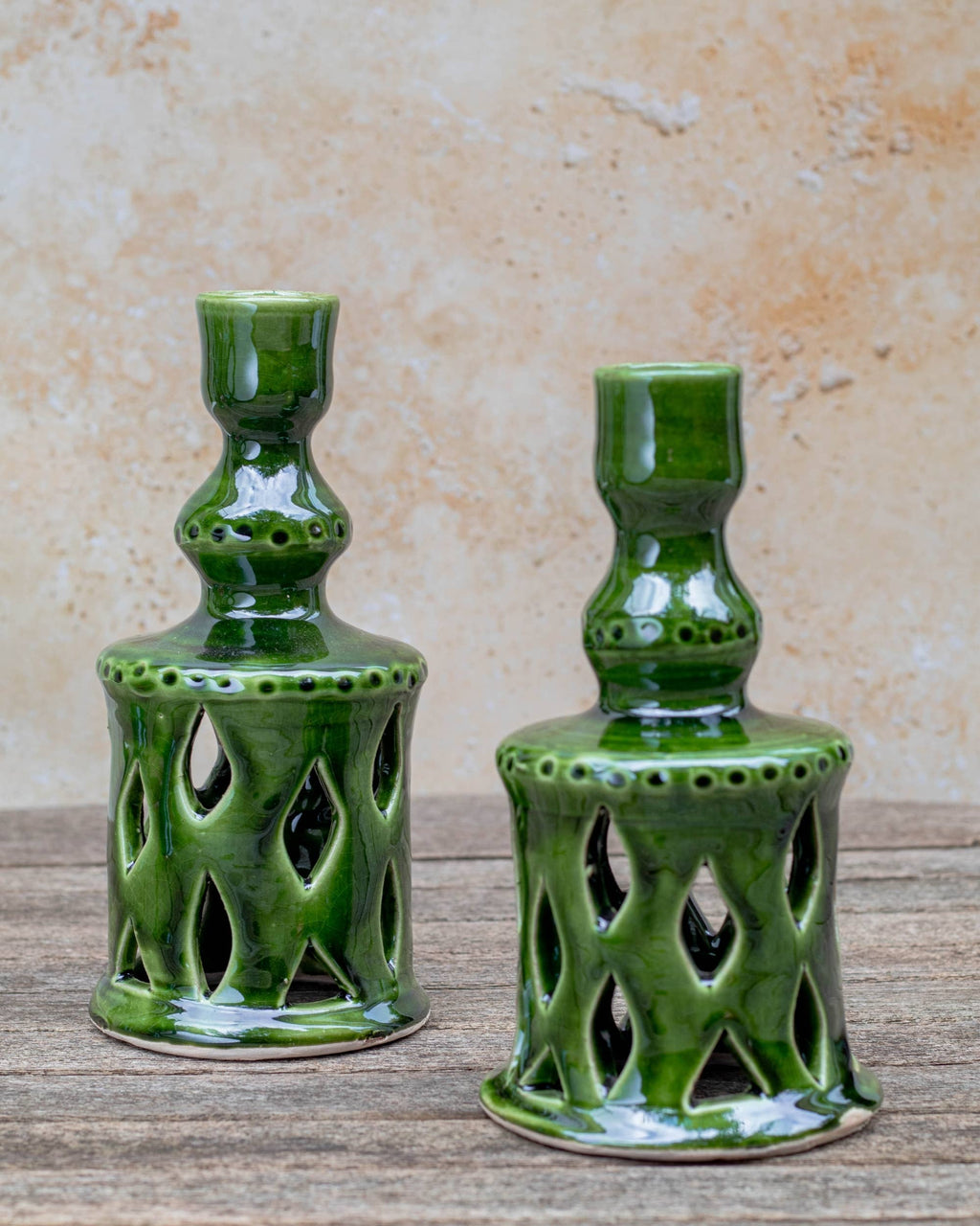 Two green glazed ceramic candle holders with openwork cutout patterns, displayed on a rustic wooden surface against a textured beige wall.