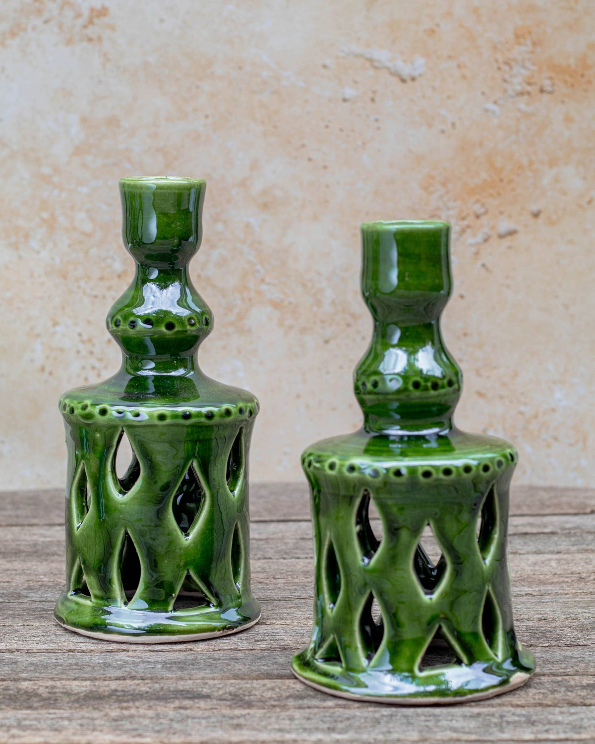 Two green glazed ceramic candle holders with openwork cutout patterns, displayed on a rustic wooden surface against a textured beige wall.