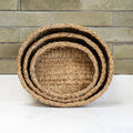 Three nested round woven baskets made of natural seagrass, stacked by size, set against a rustic brick wall and white surface.