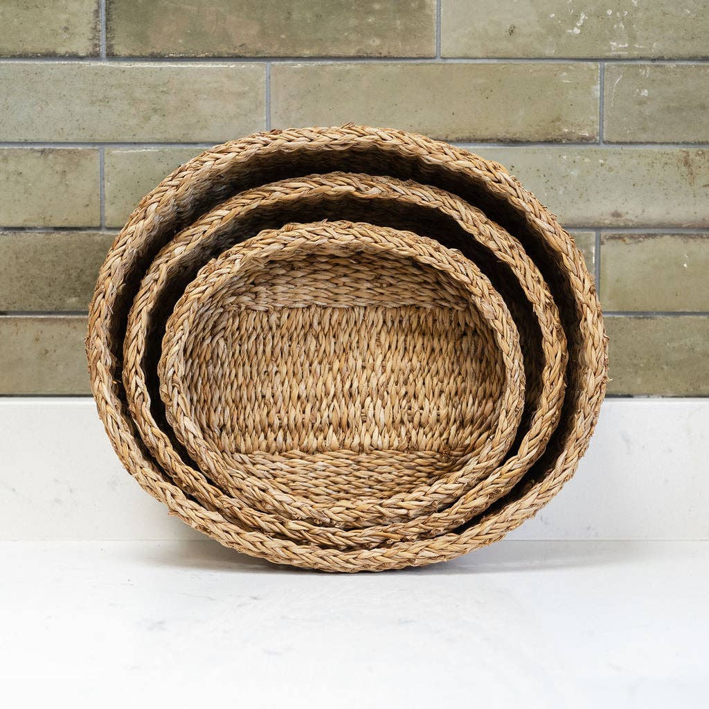 Three nested round woven baskets made of natural seagrass, stacked by size, set against a rustic brick wall and white surface.