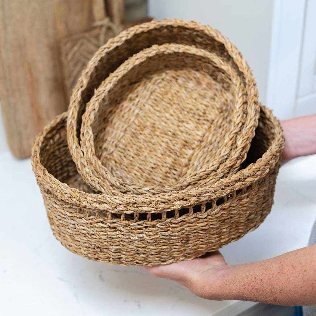Three nested round woven baskets made of natural seagrass, held by hands over a white countertop in a bright indoor setting.