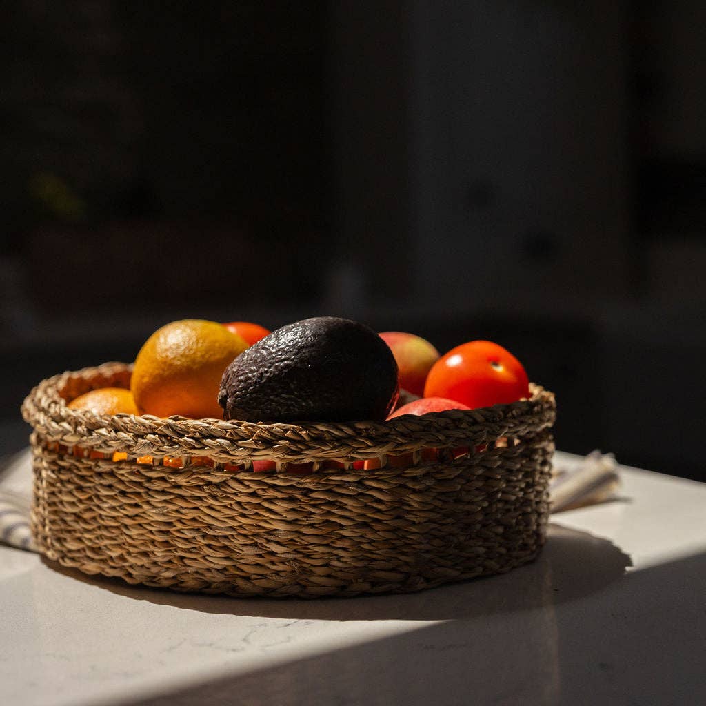 A round woven seagrass basket filled with fruit, including an avocado, orange, and tomatoes, set on a table in dramatic sunlight.