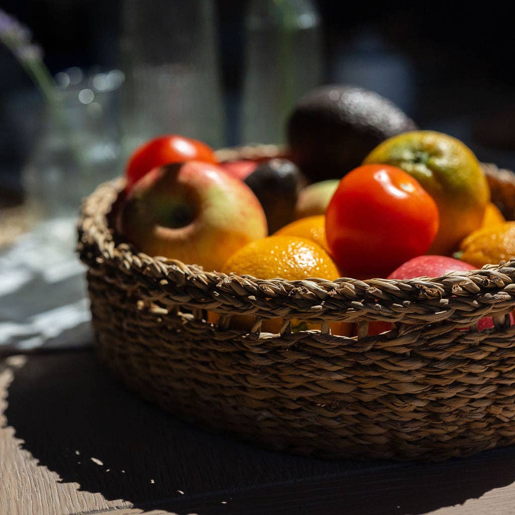 A woven oval basket filled with fresh fruit, including a red tomato, an orange, an apple, and an avocado, set on a dark wooden table in warm natural light.