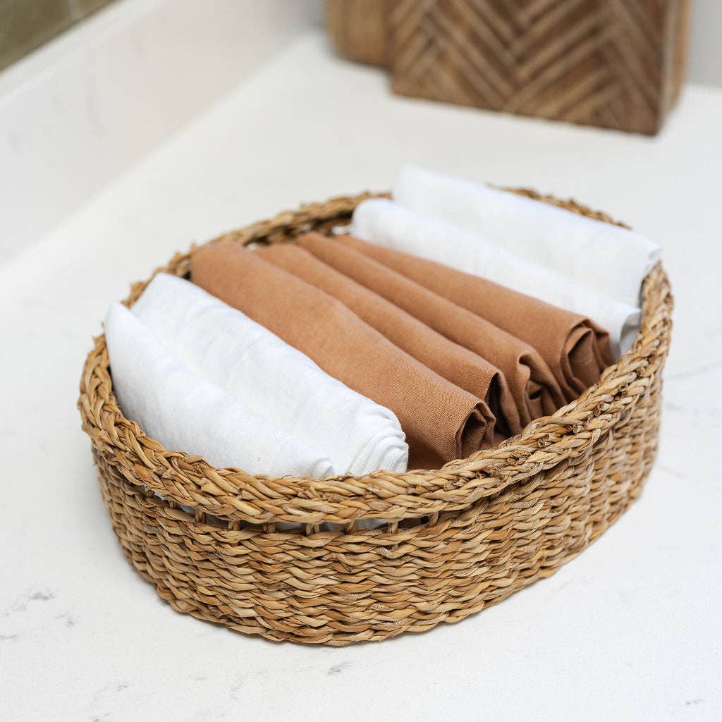 A round woven seagrass basket holding several rolled towels or cloths in white and warm tan/brown tones, placed on a light stone or marble surface.