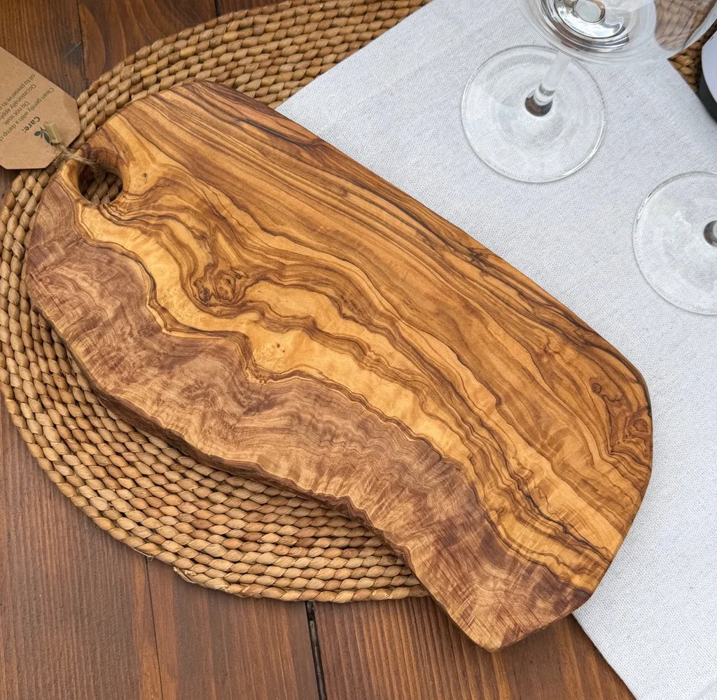 Olive wood serving board with a live edge and hanging hole on a woven placemat with wine glasses.