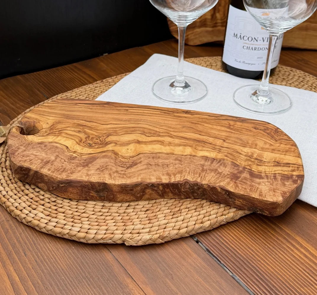 Olive wood serving board with a live edge on a woven placemat with wine glasses in the background.