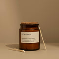 Lemongrass and May Chang natural scented candle in an amber glass jar with a cork lid and matches on a beige surface.