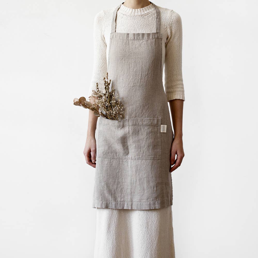 A person wearing a light grey linen apron holding a dried plant stem against a plain white background.