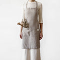 A person wearing a light grey linen apron holding a dried plant stem against a plain white background.
