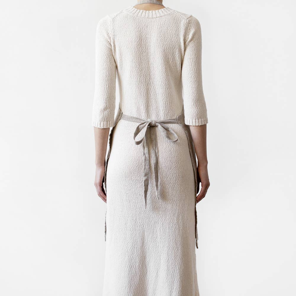 Back view of a person wearing a light gray linen apron with tied straps against a plain white background.
