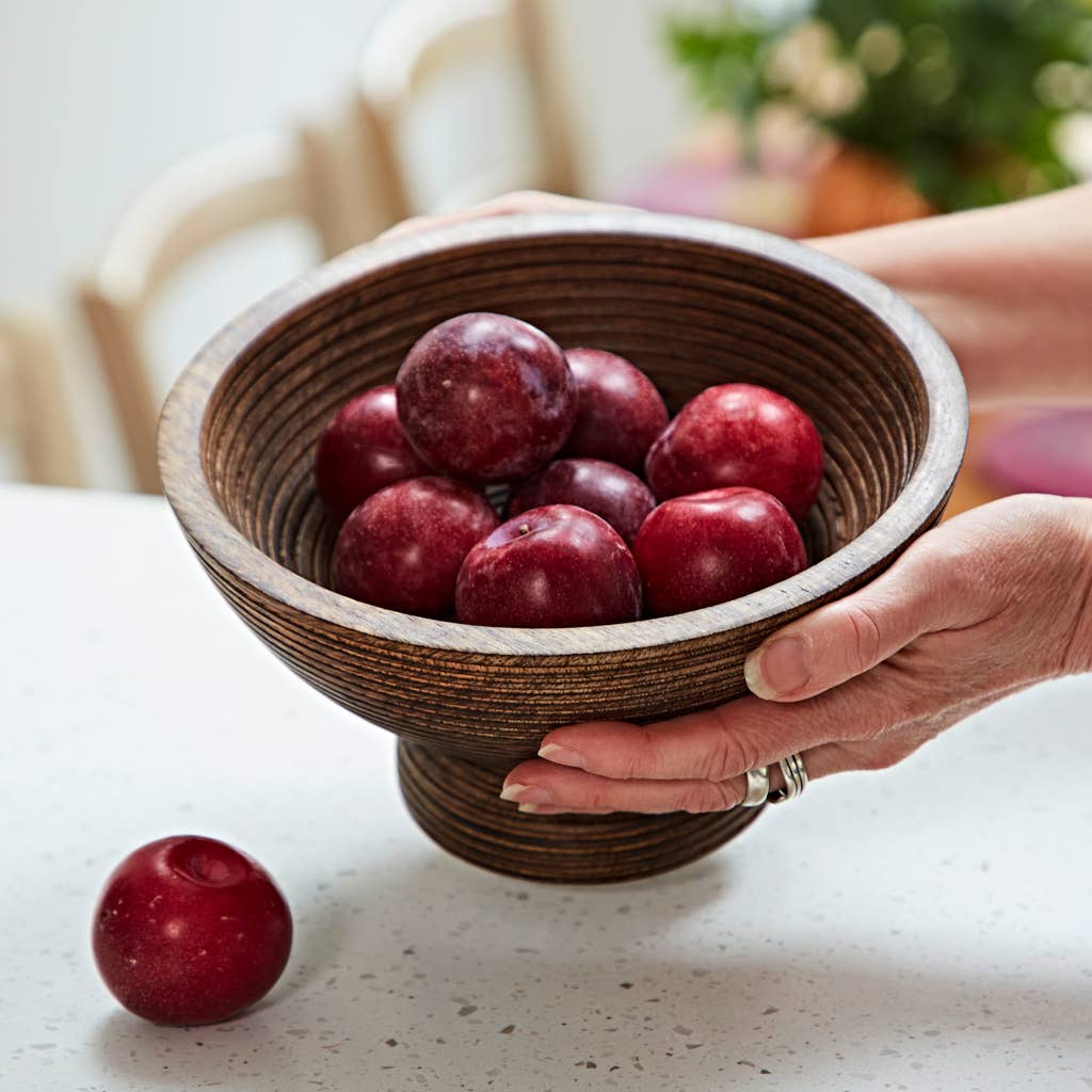 Carved mango wood pedestal bowl filled with fresh red plums over a countertop.