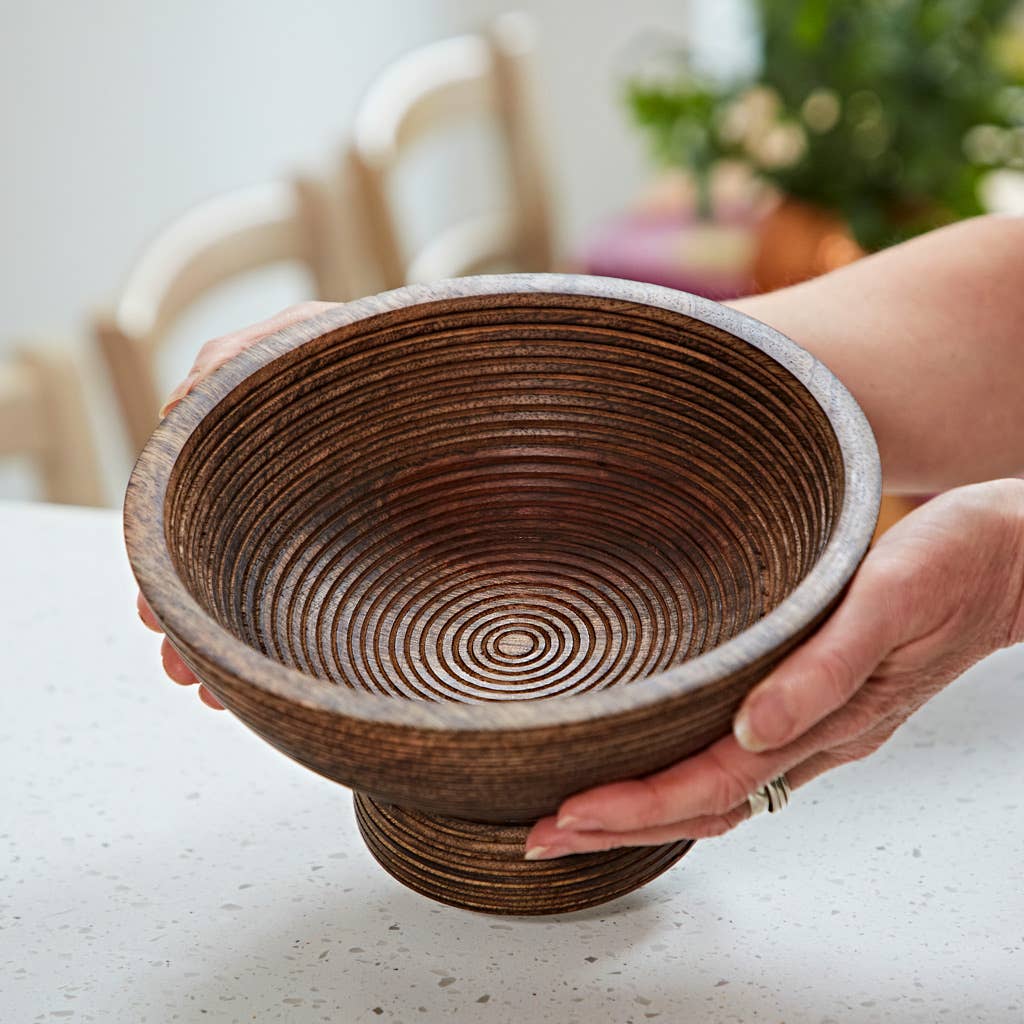 Carved mango wood pedestal bowl with spiral detail held by two hands above a table.