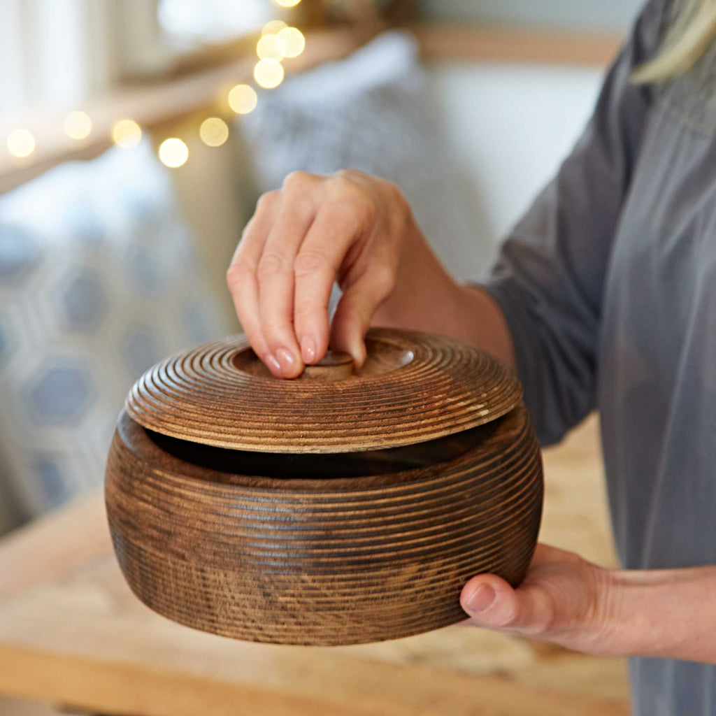 Hand holding a handmade wooden bowl with lid styled over a natural wood tabletop.