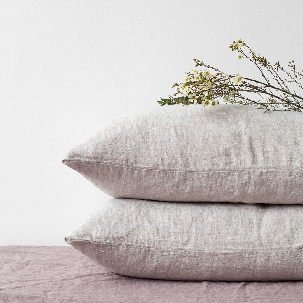 Two light gray melange linen pillowcases stacked neatly on a bed, styled with a delicate flower branch on top against a crisp white background.
