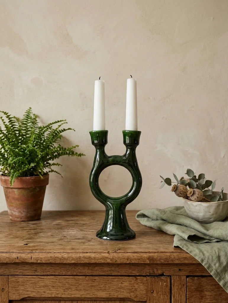 A dark green ceramic two-light candle holder with a circular center cutout, styled on a rustic wooden table with a potted fern on the left and a linen cloth with dried flowers on the right, against a textured beige wall.