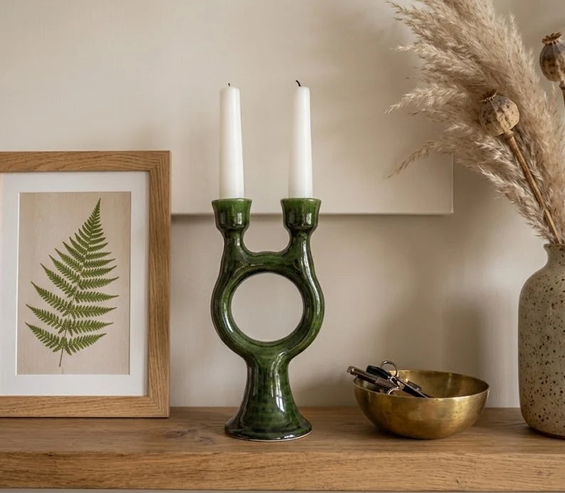 A green ceramic candle holder with two white taper candles, styled on a wooden shelf next to a framed fern print, a gold bowl, and dried pampas grass in a speckled vase.