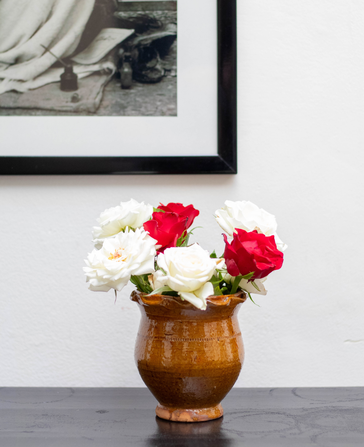 A rustic handmade terracotta plant pot filled with white and red roses, set on a dark wooden table against a white wall with a framed black-and-white photograph hanging above.