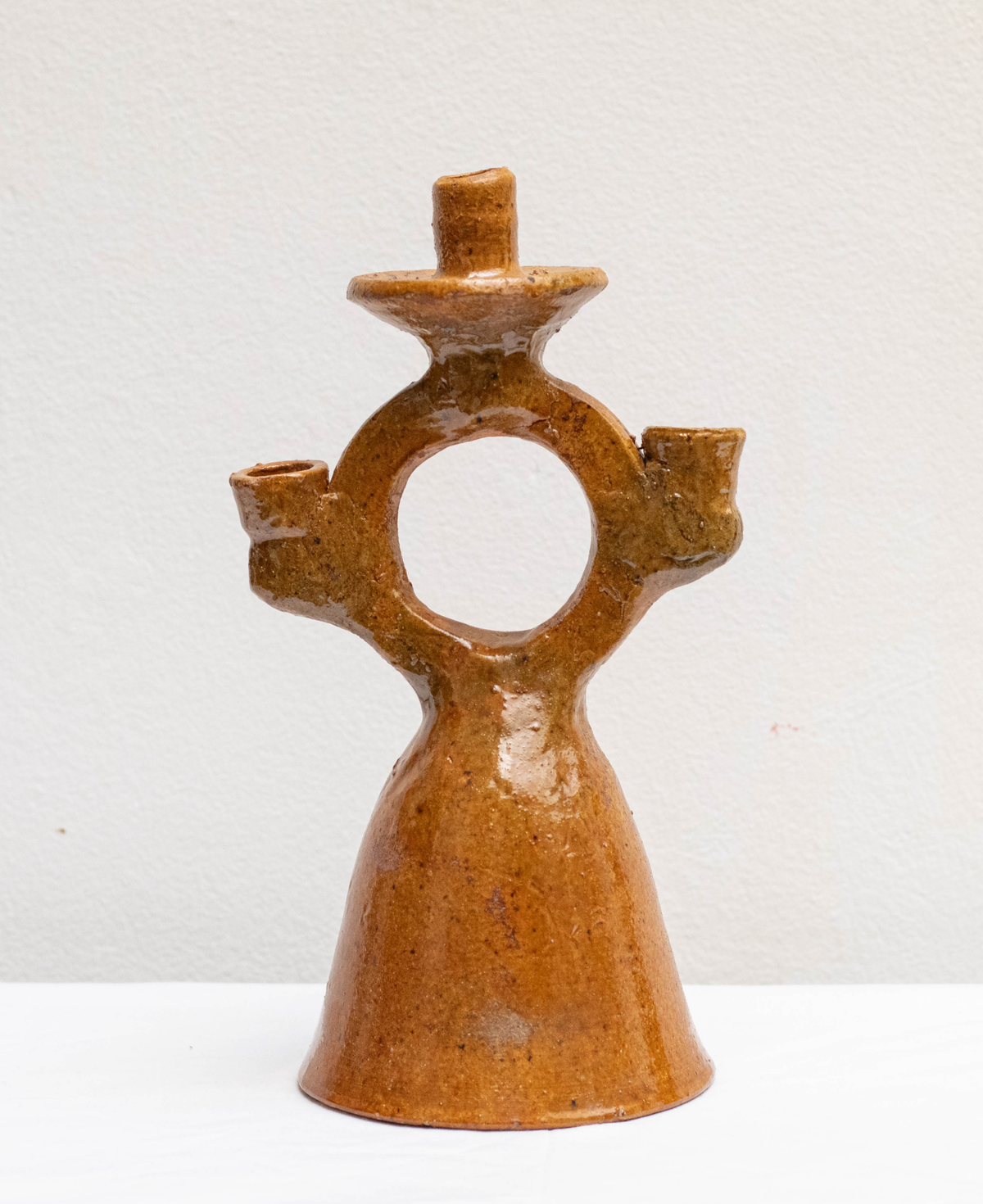 A handmade ceramic three-light candle holder with a unique ring-shaped back and a textured, glossy brown glaze, set against a plain off-white background.