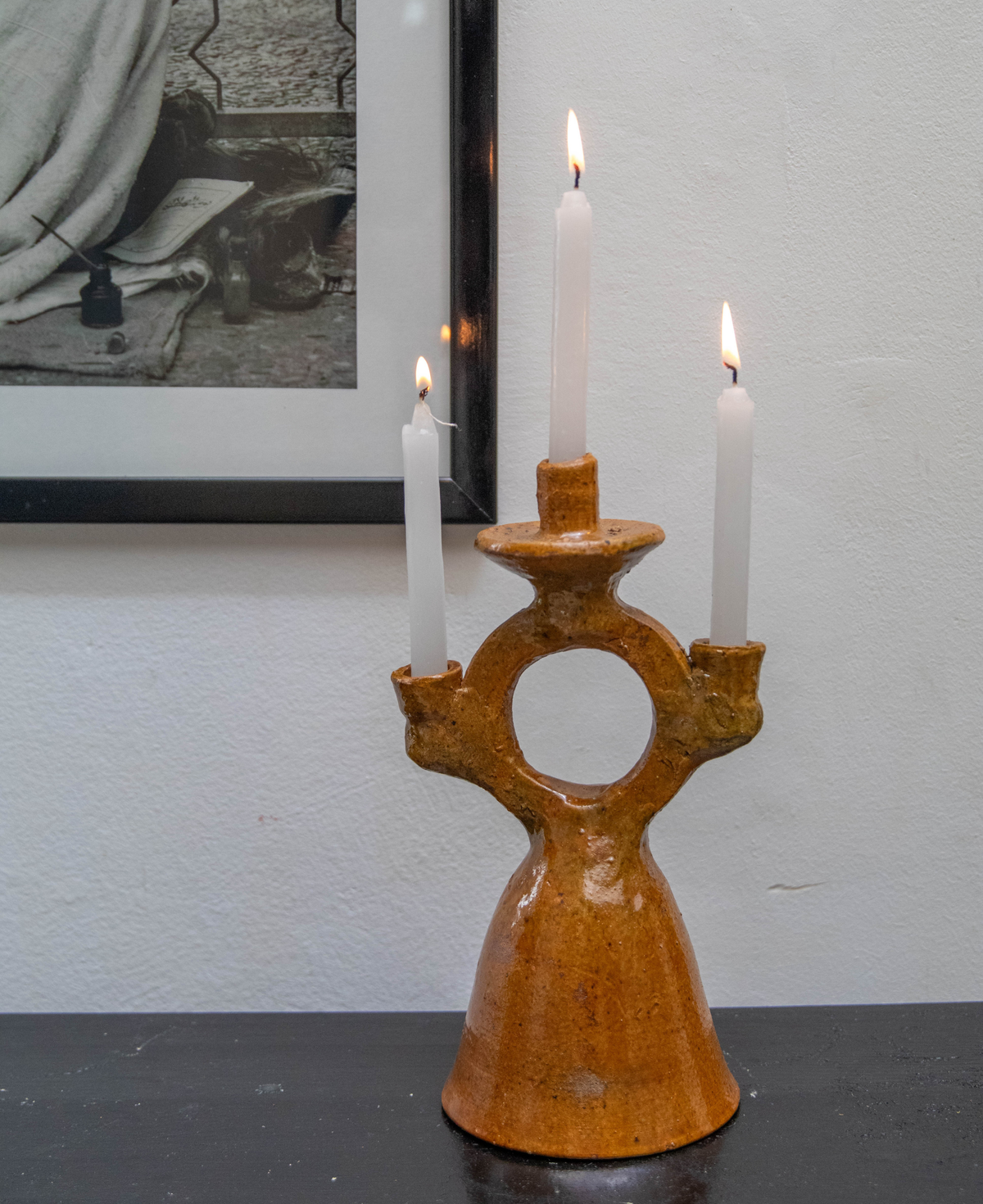 A handmade ochre terracotta three-arm candelabra holding three lit white taper candles, set on a dark tabletop against a white wall with a framed print behind it.