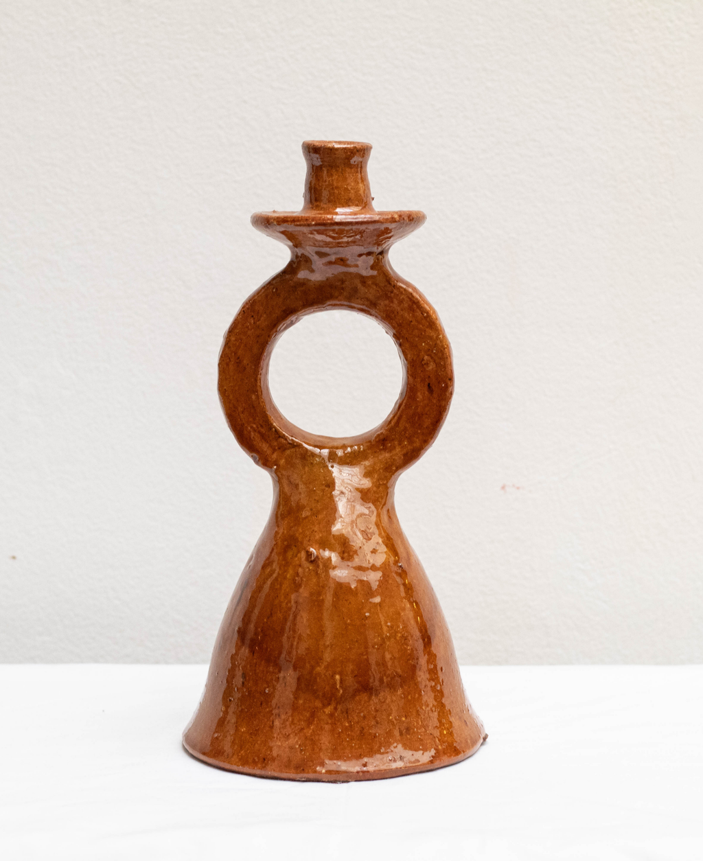 A handmade terracotta candleholder with a glossy terracotta ochre glaze, featuring a distinctive ring handle and a small top stem, set against a plain off-white background.