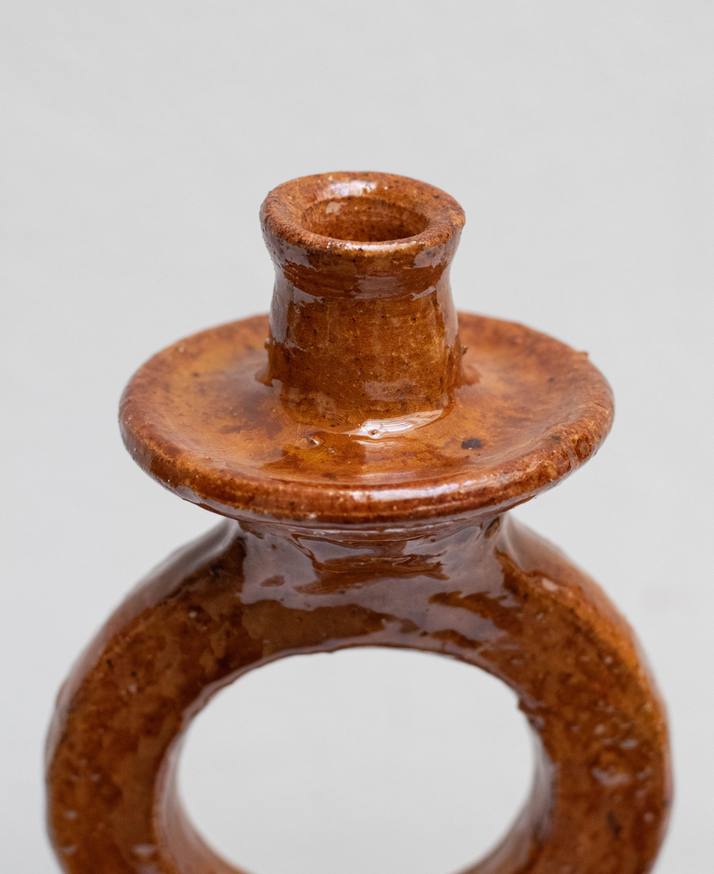 A close-up of a small glazed terracotta candle holder with a circular base and cylindrical top, shown on a plain light gray background.