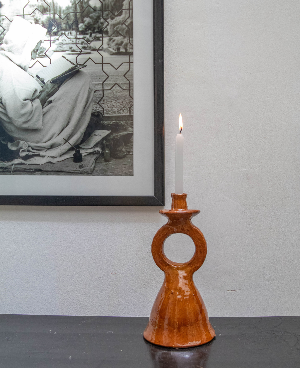 A terracotta glazed candle holder with a ring cutout, holding a lit white candle, set on a dark tabletop against a light gray wall with a framed black-and-white print in the background.