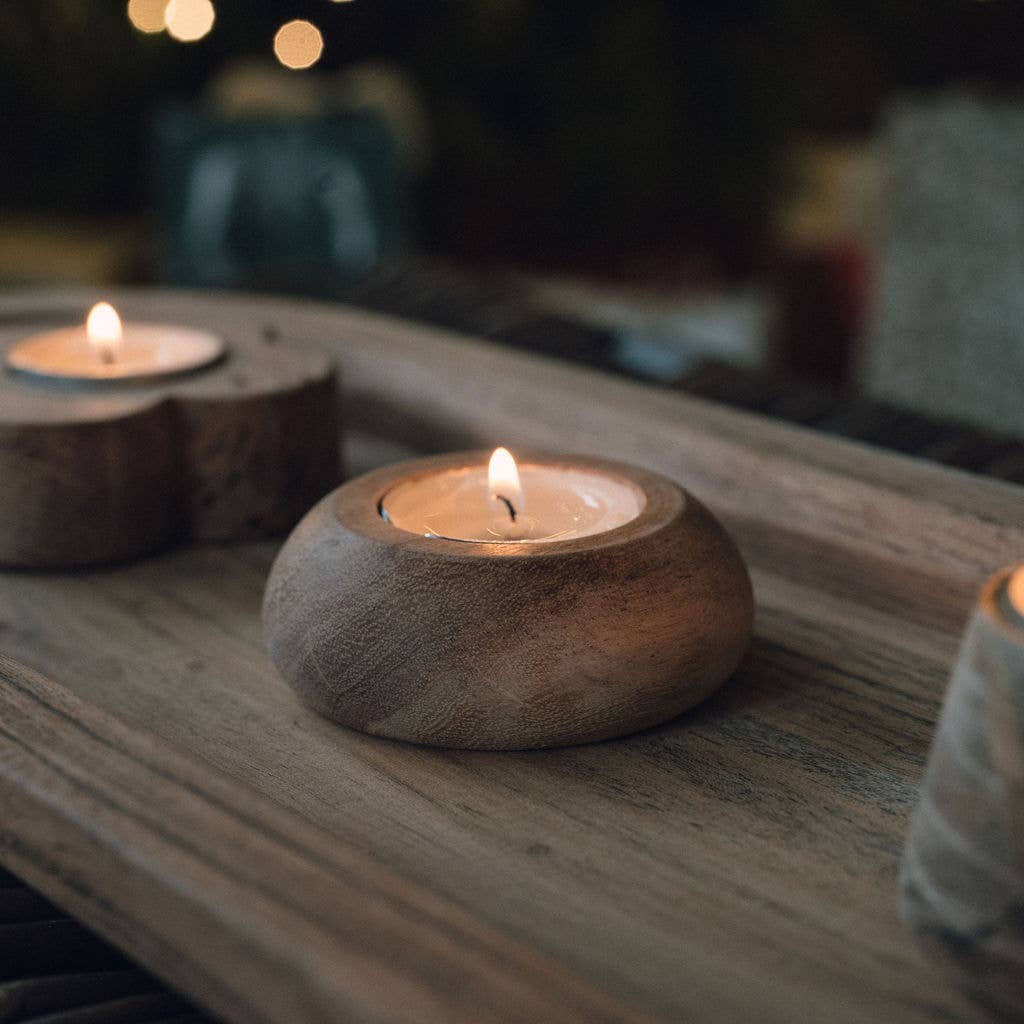 A small round wooden candle holder cradling a lit tealight candle, placed on a rustic wooden surface with other candles softly blurred in the background.